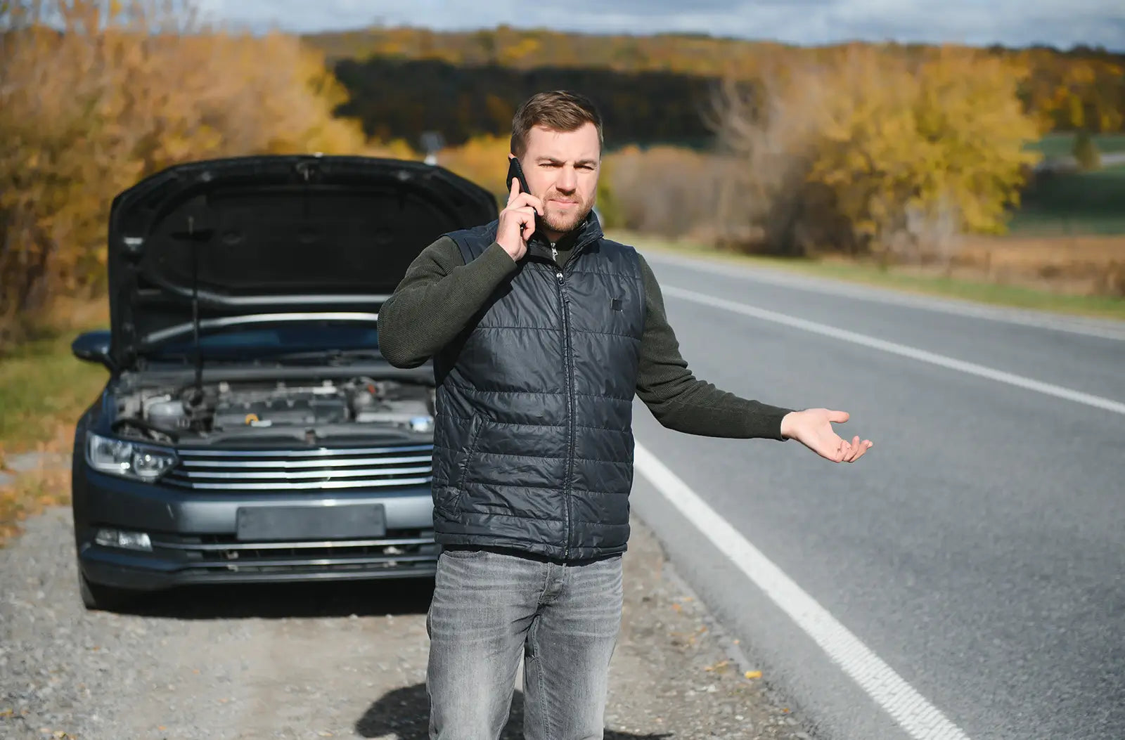Shows a frustrated male driver standing next to a dark gray car with its hood propped open by the side of a rural two-lane road, making a phone call.