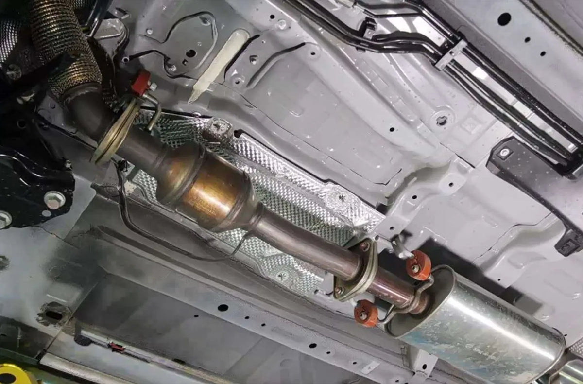 An under-car profile view of a vehicle's metallic exhaust pipe system, showcasing a threaded oxygen ($O_2$) sensor screwed directly into the catalytic converter piping.