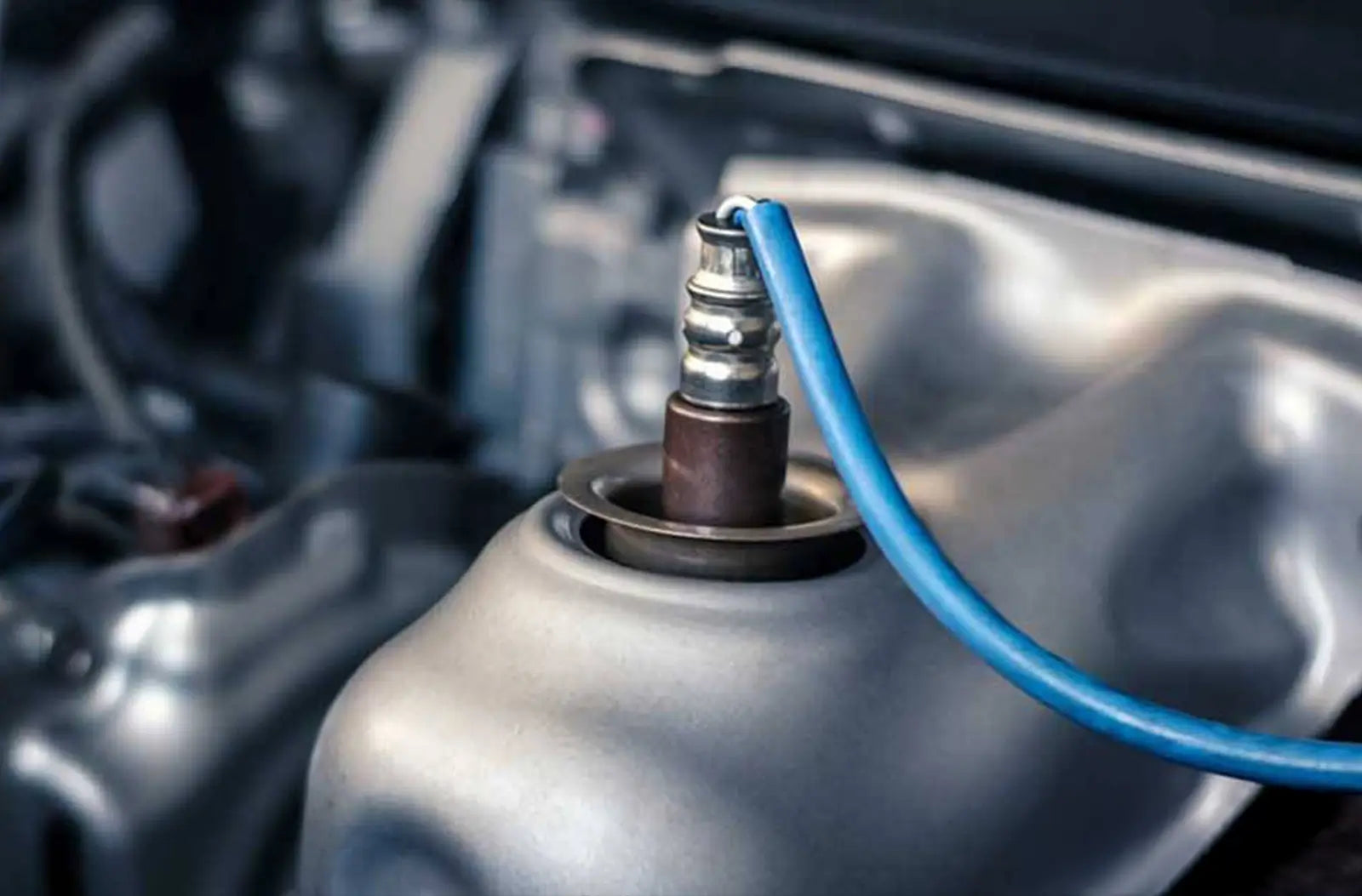 A close-up shot of a corroded, metallic oxygen ($O_2$) sensor installed directly into the exhaust pipe underneath a vehicle.