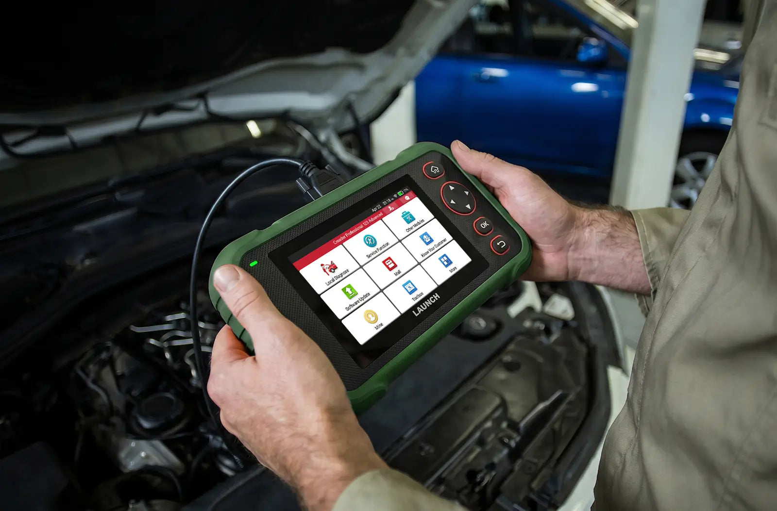 a person's hands holding a LAUNCH CRP123 Advanced diagnostic scanner over an open car engine bay in a garage setting. The device is enclosed in a rugged orange casing and features a color touchscreen displaying various automotive service menu icons.