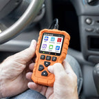 This image shows a person's hands holding and operating a Sparkace G700 diagnostic scanner inside a car. The handheld device is powered on, displaying its main menu while being used for vehicle troubleshooting.