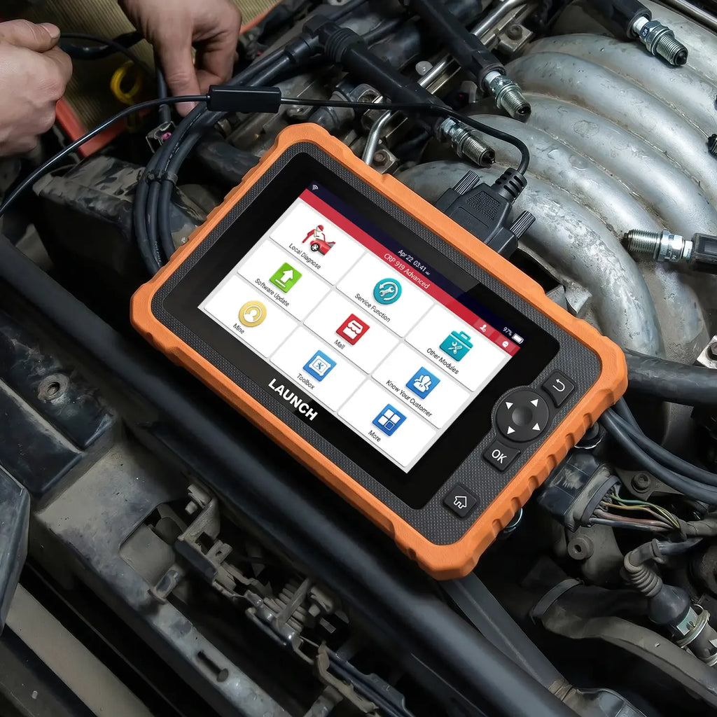 A LAUNCH CRP 919 Advanced diagnostic tablet resting on a car engine while being used for vehicle maintenance. The device features a rugged orange casing, a touchscreen with menu icons like Service Function and Local Diagnose, and is connected to the vehicle via a black OBD II cable.